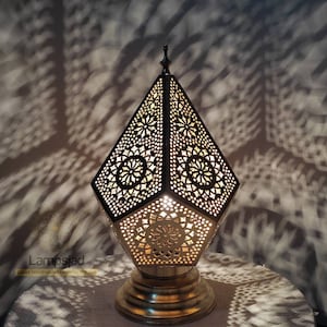 May include: A gold-toned metal table lamp with a geometric, diamond-shaped design. The lamp has intricate cut-out patterns and a decorative base.