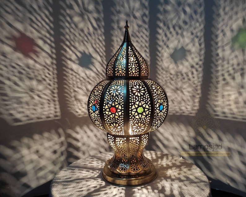 May include: A gold Moroccan table lamp with intricate cutouts and colorful glass accents. The lamp casts beautiful shadows on the wall behind it.