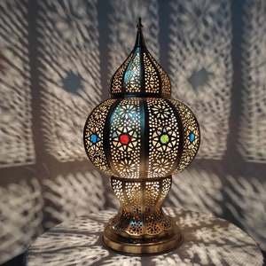 May include: A gold metal table lamp with a round, ornate base and a dome-shaped shade. The shade has a geometric pattern and features colorful glass accents.