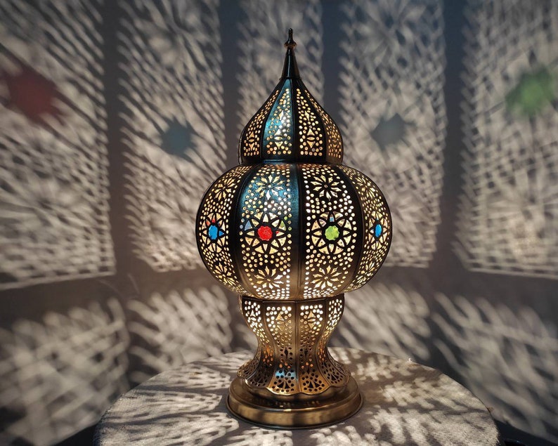 May include: A gold metal table lamp with a round, ornate base and a dome-shaped shade. The shade has a geometric pattern and features colorful glass accents.