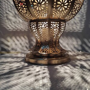 May include: A gold metal Moroccan lantern with intricate cut-out designs. The lantern has a round top and a wide base. The lantern is lit and casting shadows on the surface below.