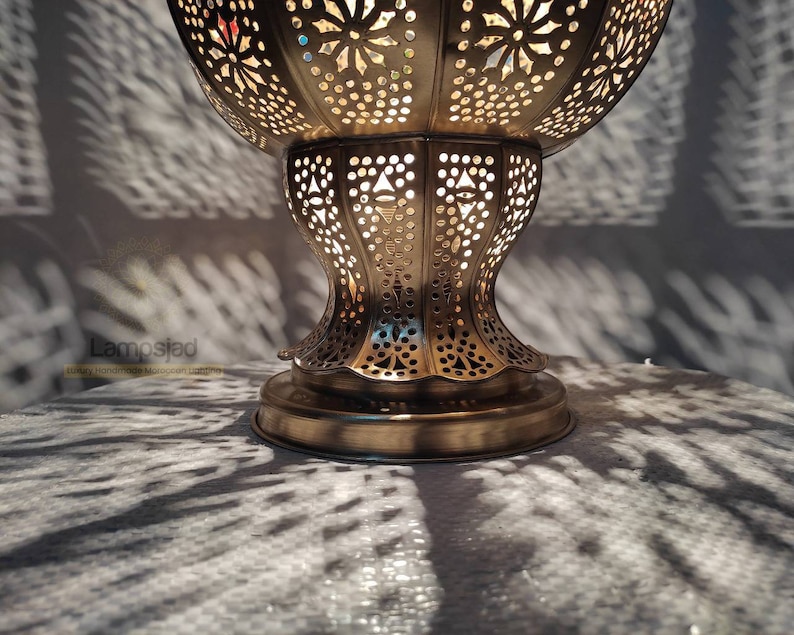 May include: A gold metal Moroccan lantern with intricate cut-out designs. The lantern has a round top and a wide base. The lantern is lit and casting shadows on the surface below.