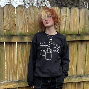 May include: Black sweatshirt with white graphic of a lighter and pipe with the text "I am not a hallucination" and "You see my pipe dream". The person is wearing black cargo pants with white stitching.