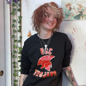 May include: Black sweatshirt with the USC Trojans logo in red and gold. The design features a Trojan helmet and the word "Trojans" in a bold font. The sweatshirt is a classic crew neck style.