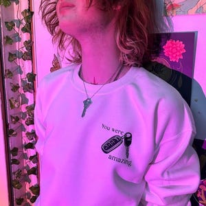 May include: White sweatshirt with the text "You were amazing" and a key graphic. The sweatshirt has long sleeves and a crew neck. A silver key pendant hangs from a necklace. The background is a pink-lit room with wall art.