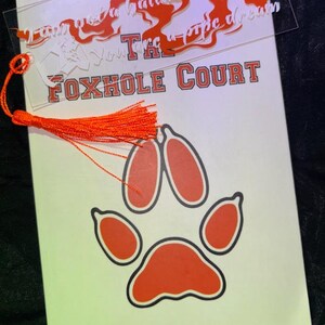 May include: A clear acrylic bookmark with an orange tassel rests on a book titled "The Foxhole Court" by Nora Sakavic. The bookmark has orange cut-out text that reads "I am not a hallucination, You're a pipe dream."