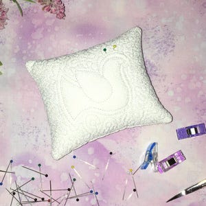 Pode incluir: Um almofadinha para alfinetes acolchoado branco com um design decorativo, rodeado por alfinetes de costura e clipes em um fundo roxo.
