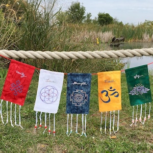 May include: A string of five colorful prayer flags with embroidered designs and words: Unity, Love, Strength, Peace, and Joy. The flags are red, white, blue, yellow, and green, with white tassels and beads. The flags are hanging on a rope.