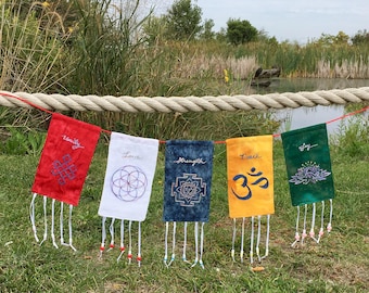 Może przedstawiać: Szereg pięciu kolorowych flag modlitewnych z haftowanymi wzorami i słowami: Jedność, Miłość, Siła, Pokój i Radość. Flagi są czerwone, białe, niebieskie, żółte i zielone, z białymi frędzlami i koralikami. Flagi wiszą na sznurku.