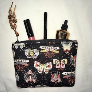 May include: A black zippered pouch featuring a repeating pattern of colorful insects and butterflies. A beaded pull tab hangs from the zipper. Inside the pouch are a pink lipstick, a black eyeliner pencil, and a gold-toned dropper bottle.