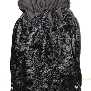 May include: Black velvet drawstring bag with a ruffled top. The bag has a textured surface and silver-colored metal hardware at the bottom. The bag is designed to be closed with a drawstring.