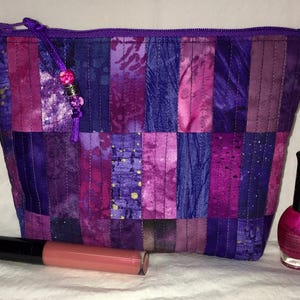 May include: A quilted cosmetic bag in shades of purple and pink, with a matching zipper and a beaded pull. A tube of lip gloss and a bottle of pink nail polish are next to the bag.