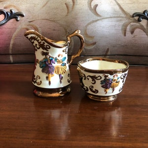May include: A white creamer and sugar bowl set with gold trim and a colourful painted design of figures in a whimsical scene.