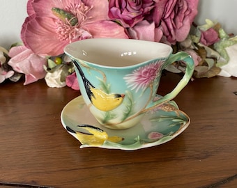 May include: A delicate porcelain teacup and saucer set featuring a floral design and two yellow birds. The cup and saucer are painted with a light blue background, green leaves, and pink flowers.