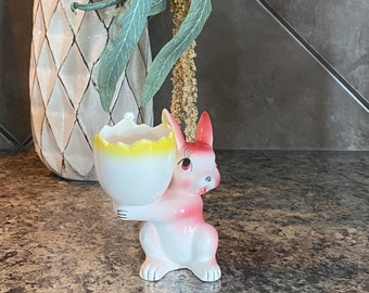May include: A white ceramic bunny figurine holding a yellow and white egg cup. The bunny has pink ears and a pink nose. The bunny is sitting on a gray countertop.