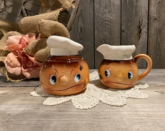 Peut inclure: Un ensemble sucrier et crémier en céramique vintage au design fantaisiste. Le sucrier et le crémier sont en forme de visages avec une peau brune, des yeux bleus et des chapeaux blancs.