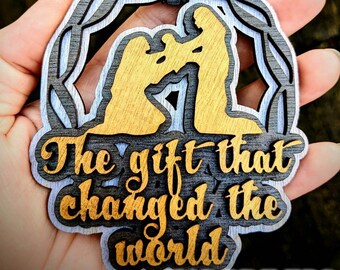 May include: A wooden Christmas ornament with a wreath design and the words "The gift that changed the world". The ornament is painted in shades of brown, black, and gold. The ornament is held in a hand.
