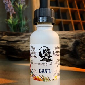 May include: A white frosted glass bottle of basil essential oil with a black dropper. The label features a wolf silhouette in a forest, the words "essential oil" and "BASIL", and floral accents. The bottle is one ounce.