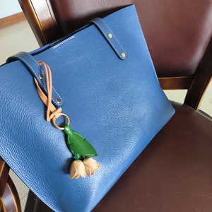 以下が含まれることがあります： 青い革製のトートバッグ。持ち手と装飾的なチャームが付いています。チャームは緑の葉と2つのベージュ色の花のつぼみです。バッグは茶色の椅子の上に置かれています。