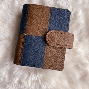 May include: A bi-fold wallet with a brown and navy blue checkerboard design. The wallet has a textured leather appearance and a brown strap closure. The background is a fluffy, white surface.
