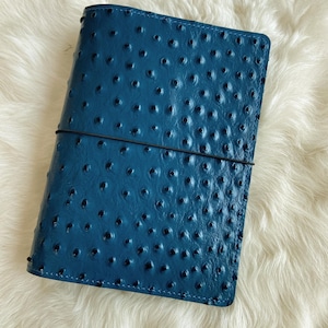 May include: A rectangular notebook cover in a deep blue color with a textured, dotted surface pattern. The cover is secured by a thin, black elastic band that wraps horizontally around the center, holding the folded leather-like material closed.