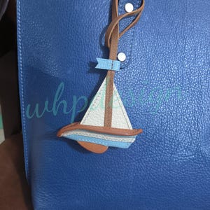 Può includere: Una borsa tote in pelle blu con un ciondolo a forma di barca a vela. Il ciondolo è realizzato in pelle bianca, marrone e blu ed è attaccato al manico della borsa con un cinturino in pelle marrone. La borsa ha una superficie strutturata.