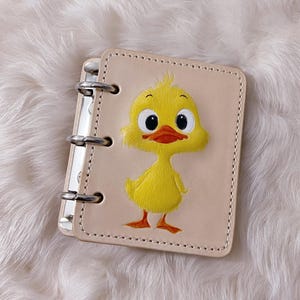 May include: A small, beige leather notebook with a metal binder. The cover features a hand-painted yellow duckling with an orange beak and feet. The notebook is open, revealing the inside pages.