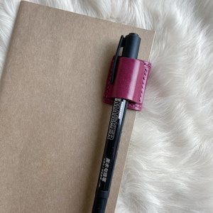 Op de afbeelding: Een zwarte permanente marker wordt vastgehouden in een bordeauxrode leren pennenhouder die aan een bruin notitieboek is bevestigd. Op de marker staat het woord "MARKER". De achtergrond is een zacht, wit oppervlak.