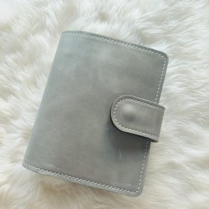 Op de afbeelding: Een grijze leren planner met een tabsluiting. De planner is rechthoekig met afgeronde hoeken en witte stiksels rond de randen. De achtergrond is een zacht, wit oppervlak.