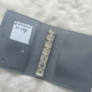 以下が含まれることがあります： 銀色のリングが付いたグレーの革製のA7リングバインダー。バインダーの前面には、「#x02006 A7 rings」と書かれた小さな白いステッカーが付いています。