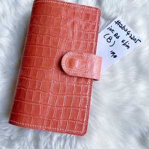 Puede incluir: Agenda de color coral con textura de piel de cocodrilo. La agenda tiene un cierre de solapa y costuras blancas. Un pequeño papel blanco con texto manuscrito está metido dentro. El texto en el papel dice "#26042205 ink B6 slim (B) 199#".
