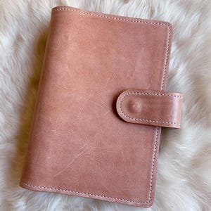 Pode incluir: Uma capa de caderno na cor rosa seco com acabamento fosco liso e costuras brancas visíveis ao longo das bordas. A capa apresenta um fecho de aba retangular no lado direito, preso com um botão de pressão circular no centro da aba.