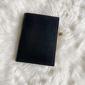 May include: A black leather wallet featuring a smooth, rectangular exterior with visible stitching along the edges. A small, gold-toned heart-shaped charm is attached to a metal zip pull on the side of the wallet, adding a subtle metallic detail.