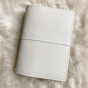 May include: A rectangular notebook cover in an off-white color featuring a textured crocodile skin pattern. The cover is secured by a thin, white elastic band that wraps horizontally around the center, holding the notebook closed.