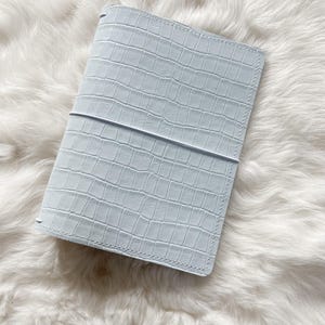 Puede incluir: Una funda de diario de cuero azul claro texturizado con un patrón de cocodrilo. La funda es rectangular con esquinas redondeadas y una banda elástica horizontal. La funda del diario está sobre una superficie blanca y esponjosa.