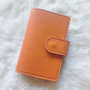 May include: An orange leather wallet with a textured surface. The wallet has a rectangular shape with rounded corners and a flap closure. The stitching is visible around the edges.
