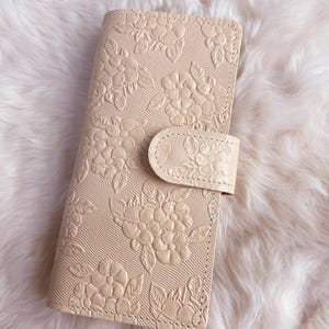 Può includere: Custodia per telefono in pelle beige con motivo floreale in rilievo. La custodia ha una chiusura a scatto e una superficie strutturata. Il motivo floreale copre l'intera parte anteriore.