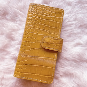 Op de afbeelding: Mosterdgele leren telefoonhoes met een krokodillenleertextuur. De hoes heeft een rechthoekige vorm met een klepsluiting en witte stiksels rond de randen. De hoes wordt getoond op een pluizig, lichtroze oppervlak.