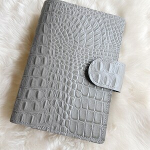 May include: A grey, faux-crocodile-skin planner with a matching tab closure. The planner is rectangular and has a textured surface. The background is a soft, white, fluffy material.