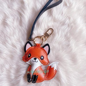 May include: A leather fox keychain in orange, white, and black with a gold-colored clasp and a dark blue wrist strap. The fox has a smiling face and is sitting upright. The keychain is on a white, fluffy surface.