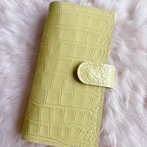 May include: A pale yellow, faux crocodile-textured planner with a snap closure. The planner is rectangular and has a stitched border. The background is a soft, white, fluffy material.