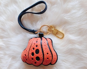 May include: A black leather keychain with a bright orange pumpkin charm. The pumpkin has a textured surface and is decorated with black circles and stitching.