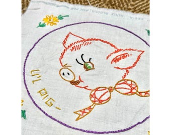 May include: Embroidered white fabric square featuring a cartoon pig with orange and red stitching. The pig is inside a purple stitched circle, with the words "Lil Pig" embroidered in yellow. Floral accents are in the corners.