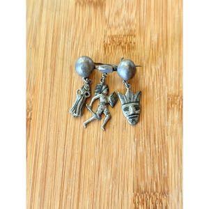 May include: A silver-toned metal brooch featuring a horizontal bar with two spherical ends. Three charms hang from the bar: a figure in a long robe, a running figure holding a shield, and a stylised mask with a feathered headdress design.