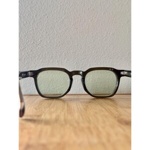 May include: A pair of dark-framed eyeglasses with square lenses. The glasses have a subtle, translucent tint and are set against a light-colored background. The frames appear to be made of a matte material.