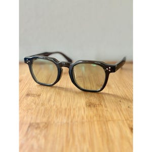 May include: A pair of dark gray eyeglasses with a square frame and round lenses. The glasses have small decorative rivets on the temples and are resting on a wooden surface. The lenses have a slight yellow tint.