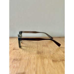 May include: A pair of eyeglasses with a dark brown frame and temples. The lenses are clear. The glasses are resting on a wooden surface. The background is a plain white wall.