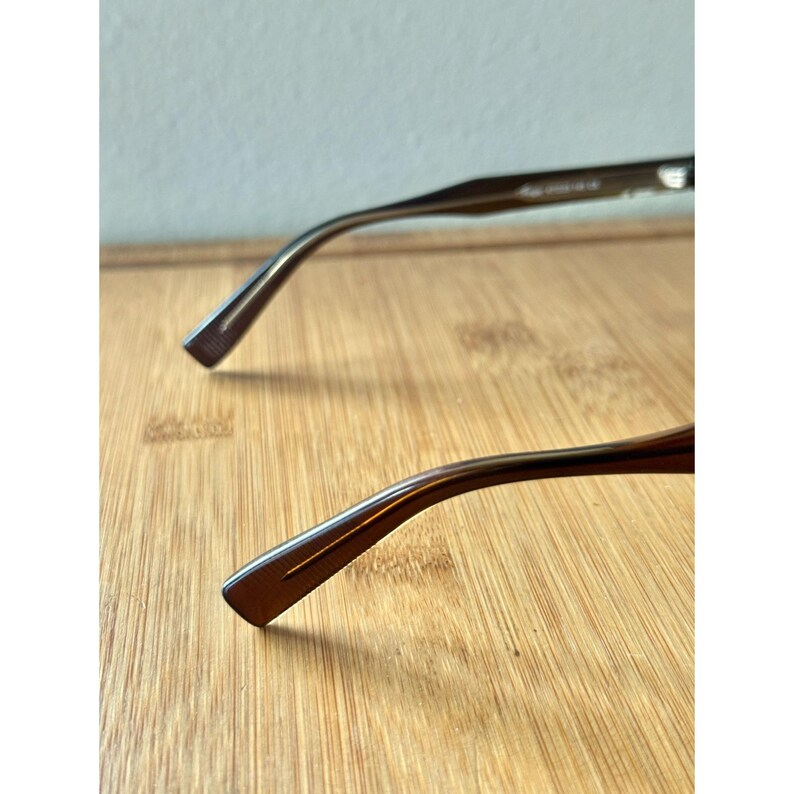 May include: Close-up of the temples of eyeglasses. The temples are a dark brown color and have a rectangular shape at the end. The background is a light brown wooden surface.