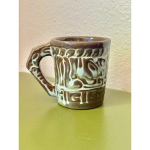以下が含まれることがあります： 茶色とオフホワイトのセラミック製マグカップ。本体の外側と持ち手には、質感のあるレリーフ状の模様が施されています。マグカップは円筒形で、厚みのある角張った持ち手が付いており、2色の釉薬の上に光沢のある仕上げが施されています。