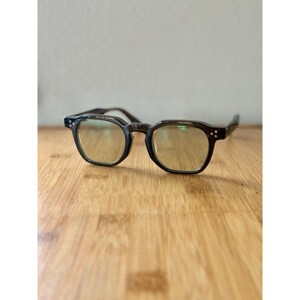 May include: Dark brown eyeglasses with a square frame and rounded edges. The lenses have a yellow tint. The glasses are set on a wooden surface.
